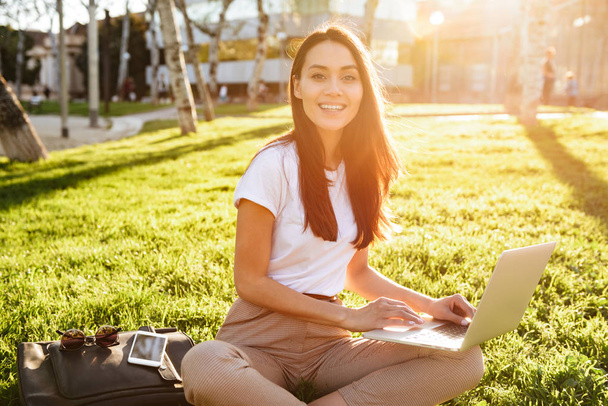 Image of amazing beautiful woman sitting on grass outdoors using laptop computer. Looking camera. - Foto, afbeelding