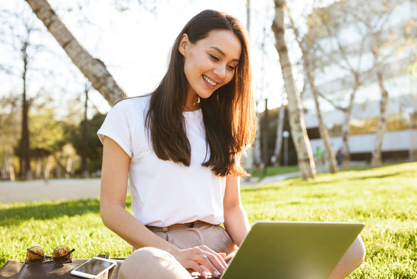 Image of gorgeous beautiful woman sitting on grass outdoors using laptop computer. Looking aside. - Foto, afbeelding