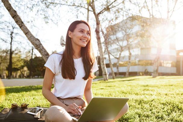 Image of gorgeous beautiful woman sitting on grass outdoors using laptop computer. Looking aside. - Foto, afbeelding