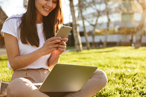 Cropped photo of woman sitting on grass outdoors using laptop computer and mobile phone. - Fotografie, Obrázek