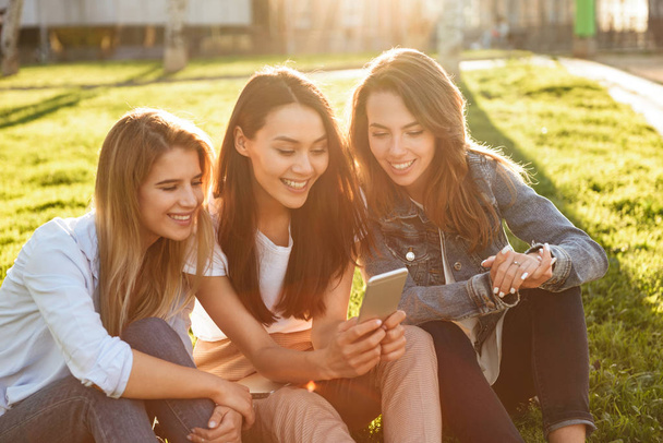 Photo of happy three friends women sitting on grass in park outdoors looking aside using mobile phone. - Фото, зображення