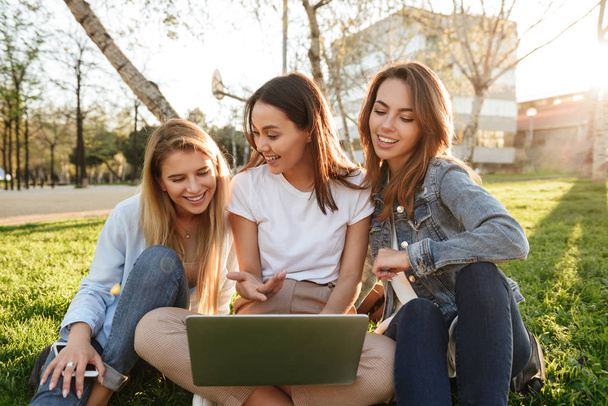Photo of amazing three friends women sitting on grass in park outdoors looking aside using laptop computer. - Foto, Bild