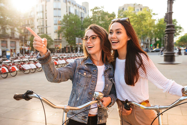Image of two young beautiful women friends outdoors on bicycles on the street pointing. - Photo, Image