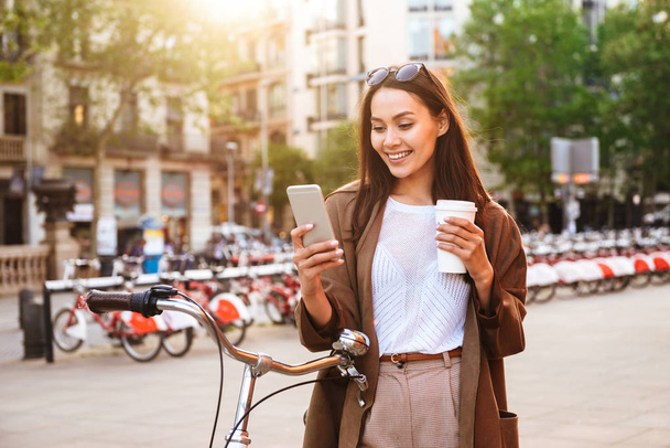 Image of young woman outdoors with bicycle on the street looking aside chatting by mobile phone drinking coffee. - 写真・画像