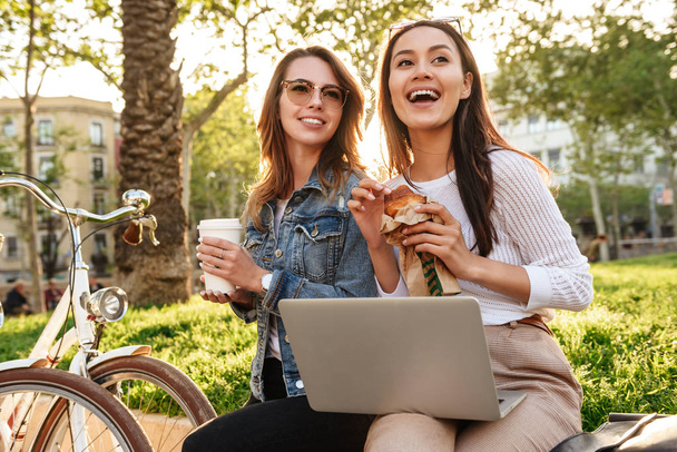 Photo of two young happy women friends outdoors with bicycles in park drinking coffee eating croissant while using laptop computer. - Photo, image