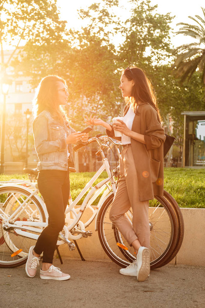 Image of two young beautiful women friends outdoors with bicycles in park using mobile phone. - Foto, Bild