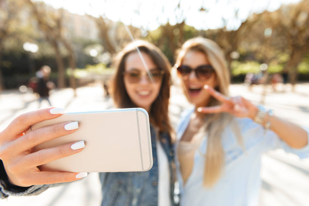 Image of two amazing smiling women friends walking outdoors make selfie by mobile phone. - Фото, изображение