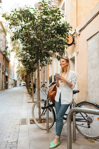 Image of amazing happy beautiful woman walking outdoors on bicycle using mobile phone - Photo, image