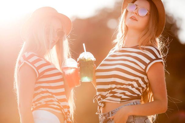Portrait of two young beautiful girls in trendy stripe shirt, beach hat, sunglasses an jeans shirt on the sea. two friends having fun at the sea with cocktails on their hands. Youth and happiness concept. - Photo, Image