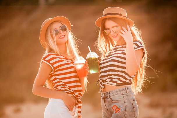 Portrait of two young beautiful girls in trendy stripe shirt, beach hat, sunglasses an jeans shirt on the sea. two friends having fun at the sea with cocktails on their hands. Youth and happiness concept. - Photo, image
