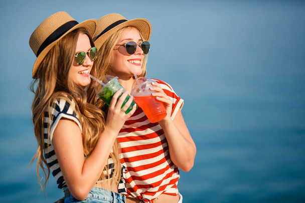 Portrait of two young beautiful girls in trendy stripe shirt, beach hat, sunglasses an jeans shirt on the sea. two friends having fun at the sea with cocktails on their hands. Youth and happiness concept. - Foto, imagen