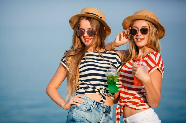 Portrait of two young beautiful girls in trendy stripe shirt, beach hat, sunglasses an jeans shirt on the sea. two friends having fun at the sea with cocktails on their hands. Youth and happiness concept. - 写真・画像
