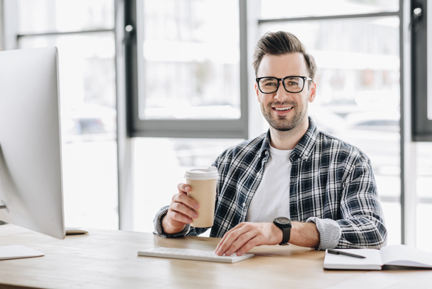 guapo joven programador sosteniendo taza de papel y sonriendo a la cámara mientras está sentado en el lugar de trabajo
 - Foto, Imagen