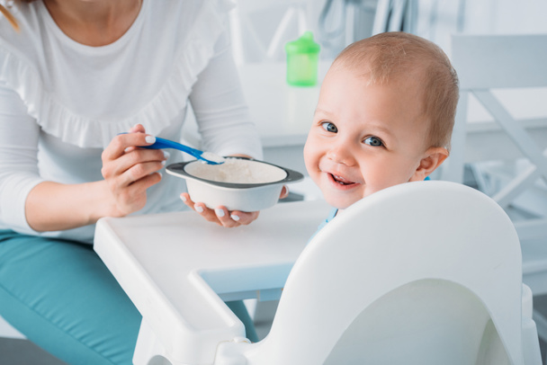 cropped shot of mother feeding happy child with porridge while he looking at camera - Foto, imagen