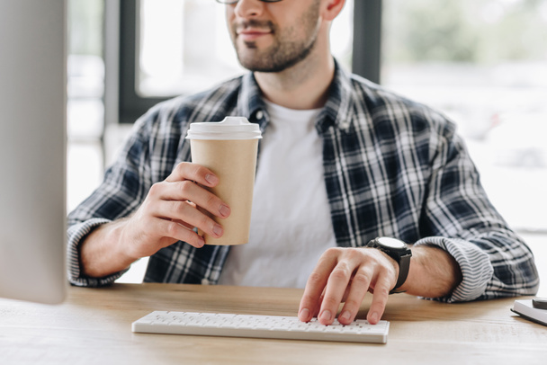 cropped shot of man holding paper cup and using desktop computer at workplace - Photo, Image