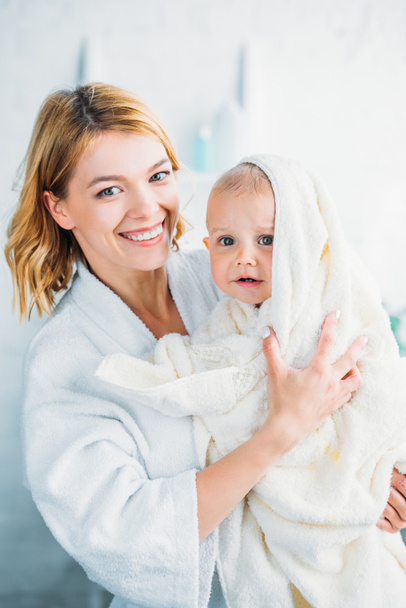 smiling mother in bathrobe holding confused little child covered with towel and looking at camera - Photo, Image