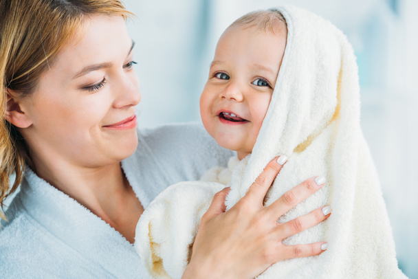 mosolygó anya, a gazdaság boldog kis gyermek fürdőköpeny törülközőt borított - Fotó, kép