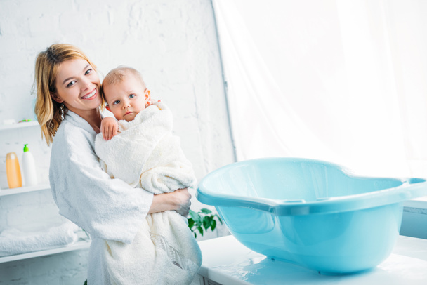 Mère souriante en peignoir portant adorable enfant couvert de serviette près de la baignoire en plastique pour bébé
 - Photo, image