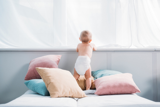 枕の多くが付いているベッドの上に立って、窓を通して見るおむつで幸せな赤ちゃん - 写真・画像