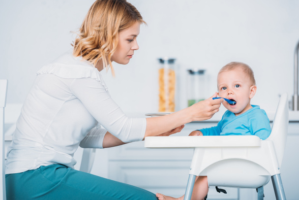 beautiful young mother feeding child while he sitting in high chair at kitchen - Фото, зображення