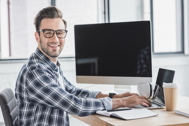 handsome young programmer in eyeglasses smiling at camera while working with laptop and desktop computer - Photo, Image