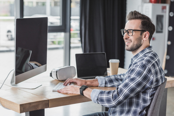 giovane uomo sorridente in occhiali da vista utilizzando computer desktop e laptop sul posto di lavoro
 - Foto, immagini