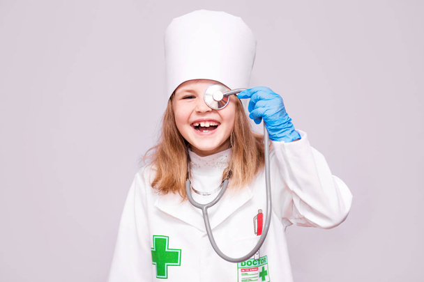 Usměvavá holčička v lékařských uniformě drží stetoskop a při pohledu na fotoaparát izolované na bílém - Fotografie, Obrázek