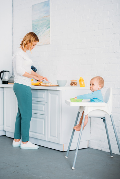 madre preparando la cena para su hijo pequeño mientras él sentado en la silla de alimentación en la cocina
 - Foto, Imagen