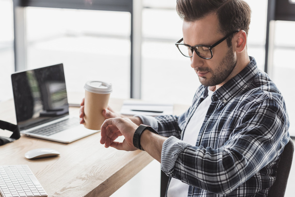 young man in eyeglasses holding coffee to go and checking smartwatch at workplace - Foto, Bild