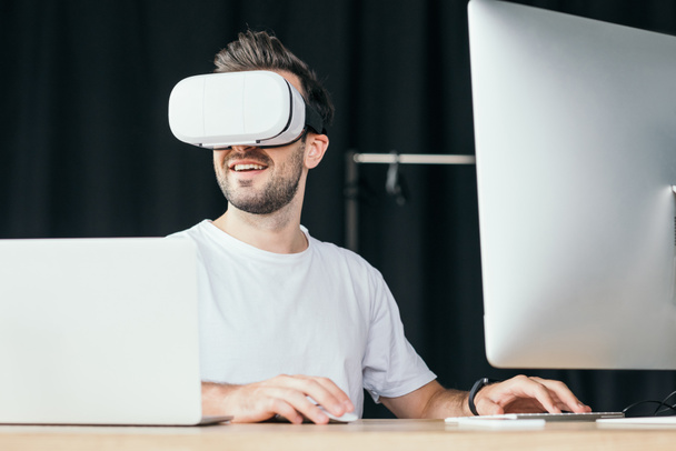 joven sonriente en auriculares de realidad virtual usando computadora de escritorio y computadora portátil
  - Foto, imagen