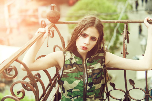 fashion makeup. Pretty girl in camouflage vest holding metallic balcony rails - Foto, afbeelding