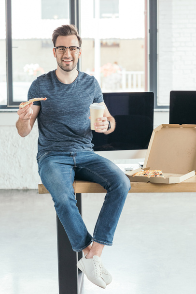 όμορφος χαμογελαστός νεαρός προγραμματιστής σε γυαλιά κρατώντας χαρτί Κύπελλο και τρώγοντας πίτσα στο χώρο εργασίας  - Φωτογραφία, εικόνα