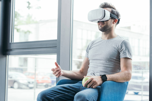 jovem sorridente em realidade virtual headset segurando maçã e sentado na cadeira
  - Foto, Imagem