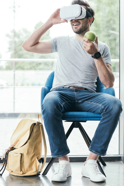 uśmiechający się młody człowiek trzyma jabłko i przy użyciu zestaw wirtualnej rzeczywistości  - Zdjęcie, obraz