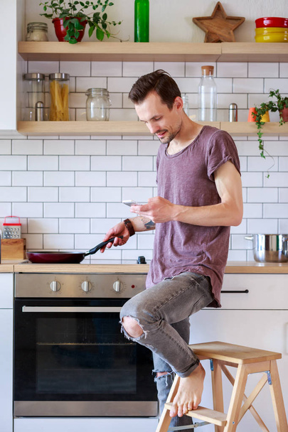 Obraz człowieka brunet z patelni i telefon w ręce w kuchni - Zdjęcie, obraz