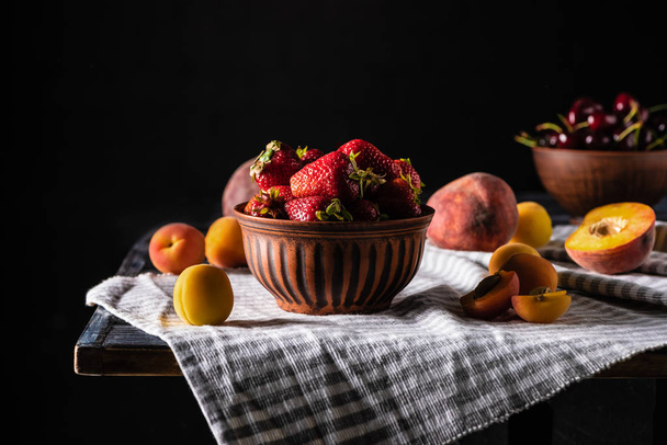 selective focus of bowl with strawberries surrounded by peaches and apricots on wooden table on black - 写真・画像
