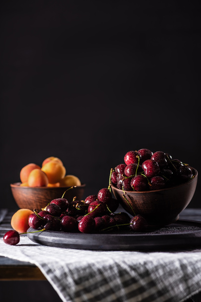 selective focus of bowls with ripe cherries and apricots on wooden table on black  - Foto, Imagem