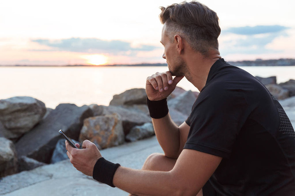 Πορτρέτο του ένα ισχυρό αθλητή χρησιμοποιώντας το κινητό τηλέφωνο, ενώ κάθεται στην παραλία - Φωτογραφία, εικόνα