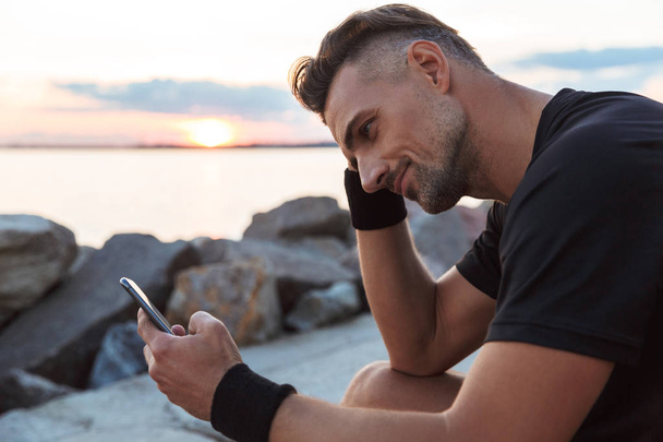 Portrait of a fit sportsman using mobile phone while sitting at the beach - Photo, image