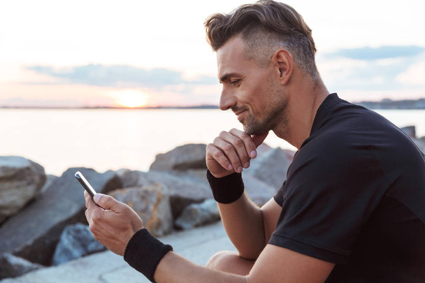Portrait of a smiling sportsman using mobile phone while sitting at the beach - Фото, изображение
