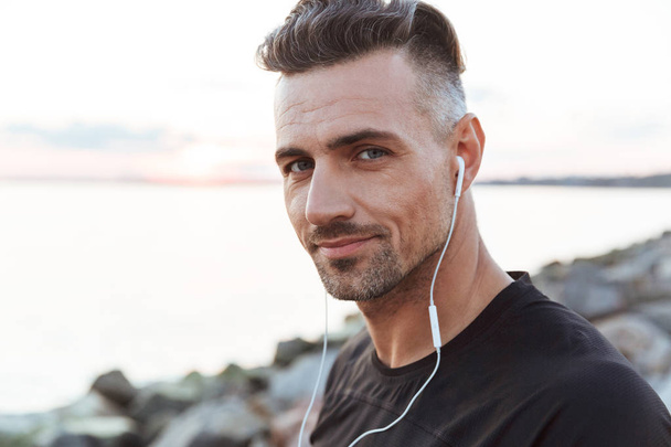 Close up portrait of a smiling sportsman listening to music with earphones while standing at the beach - Fotó, kép