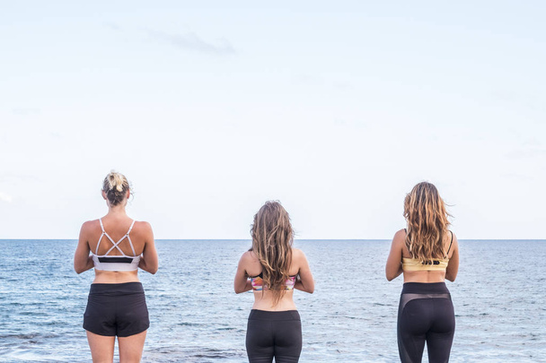 three beautiful girls viewed from back side to yoga meditation fitness workout in front of the ocean to take energy of nature and enjoy a freedom alternative lifestyle. outdoor leisure sport activity concept - Photo, Image
