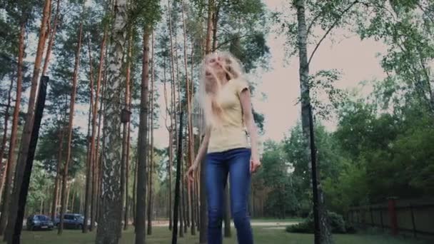 Joyeux saut féminin sur trampoline. Belle jeune femme blonde pieds nus en chemise d'été légère et jeans regardant la caméra et souriant sautant sur un grand trampoline avec de grands arbres autour
. - Séquence, vidéo