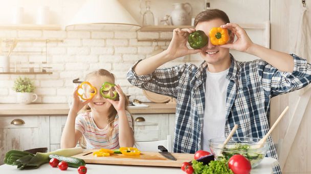 Happy father and his cute daughter covering eyes with pepper, having fun while they are making healthy meal together at kitchen, copy space - Photo, image