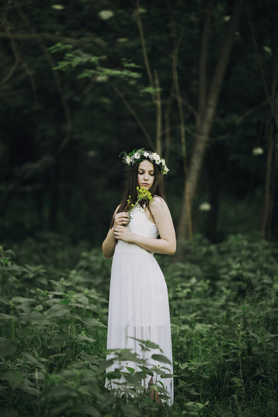 Outdoor portrait of beautiful young woman in a white dress posing behind wild flowers - Fotó, kép