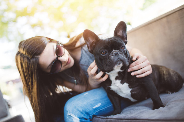 Cute teenager girl enjoying outdoors in cafe bar together with her adorable French bulldog puppy. Strong back light. Warm sunny day. - Photo, image