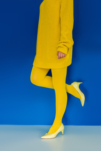 Обрізаний вид дівчини в жовтому одязі та взутті на каблуці на синьому фоні
 - Фото, зображення