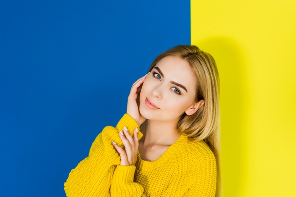 Krásná blond dívka, která nosí bílý svetr na modré a žluté pozadí - Fotografie, Obrázek