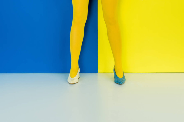 Ausgeschnittene Ansicht weiblicher Beine in verschiedenen Schuhen auf blauem und gelbem Hintergrund - Foto, Bild