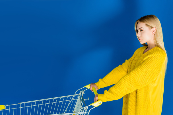 attraktive junge Mädchen schieben Einkaufswagen auf blauem Hintergrund - Foto, Bild
